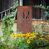 A decorative metal screen with a floral design stands in a garden, surrounded by yellow flowers and greenery, with a rustic wooden building in the background. Produktbild-Vorschau