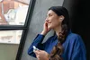 A woman is looking out a window while holding a white charging case and wearing a silver earbud. Anteprima dell’immagine prodotto