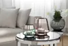 Two glass candle holders with lit candles on a reflective table, with a sofa and flowers in the background. Produktbild-Vorschau
