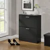 A black shoe cabinet with two flip drawers stands in a room next to a window. A pair of black high-heeled shoes are on the floor in front of the cabinet. Náhled obrázku produktu