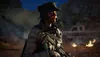 A soldier in tactical gear stands in a desert environment at dusk, with a pickup truck and fires in the background. Produktbild-Vorschau