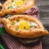 Two khachapuri (Georgian cheese bread with egg) are presented on a wooden table, one in the foreground and another in the background, both on a red and white checkered cloth. Produktbild-Vorschau