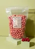 A large bag of pink raspberry crispy balls sits on a light green surface, with a few fresh raspberries in the foreground. Produktbild-Vorschau
