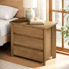 A wooden nightstand with three drawers is placed next to a bed, with a lamp and books on top. A window with greenery is visible in the background. Produktbild-Vorschau