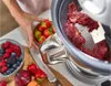A close-up shot of a food mill with berries and other fruits nearby, and a person's hands reaching for a bowl of strawberries. Produktbild-Vorschau