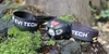 A black and gray LED headlamp with a black strap is resting on mossy rocks and tree stumps outdoors. Náhled obrázku produktu