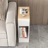 A narrow side table with a drawer and shelf is placed next to a sofa, with books on the shelf and a magazine and coffee cup on top. Produktbild-Vorschau