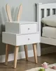 A white bedside table with two drawers and wooden legs, decorated with wooden bunny ears, is placed next to a white bed. A small toy tea set is visible on the floor in the foreground. Náhľad obrázku produktu