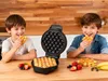 Two boys are in a kitchen, one eating a waffle and the other holding one, with a waffle maker and fruit on the counter. Náhľad obrázku produktu