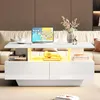 A modern white coffee table with LED lighting in a living room setting, featuring a tablet, headphones, books, and a vase of flowers. Produktbild-Vorschau