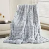 A light gray plush blanket draped over a chair, with a side table and decorative items in the background. Náhled obrázku produktu