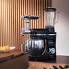 A black and silver multi-functional kitchen appliance, including a mixer, meat grinder, and blender, is displayed on a dark countertop. The background features vertical wooden slats. Náhled obrázku produktu