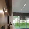 A modern living room with a track light on the ceiling and a wall lamp, overlooking a garden through large windows. Produktbild-Vorschau