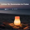 A glowing lantern sits on a sandy beach at dusk, with the ocean and distant lights in the background. Produktbild-Vorschau