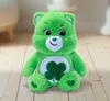 A green stuffed bear with a shamrock on its belly sits on a wooden floor. Produktbild-Vorschau