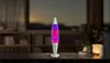 A lava lamp is placed on a table in front of a blurred background of a room with shelves and a window. Náhľad obrázku produktu