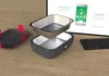 A heated lunchbox and its components are displayed on a wooden surface, with a tablet showing a smartphone app interface in the background. Produktbild-Vorschau