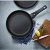 A frying pan is shown from a top-down perspective on a textured gray surface, with a wooden cutting board, a lemon wedge, and rosemary sprigs to the side. Produktbild-Vorschau