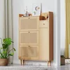 A light wood shoe cabinet with rattan detailing, placed in a room with a plant and curtains. Produktbild-Vorschau