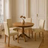 A dining set with a round wooden table and four cream-colored chairs in a room with white paneled walls and a large window. Produktbild-Vorschau