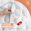 A close-up of a white mattress topper with a hand demonstrating an elastic strap. Produktbild-Vorschau
