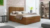 A bedroom scene featuring a brown boxspring bed with pillows and a blanket, a side table with a lamp, and a bookshelf in the background. Náhled obrázku produktu