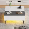 A modern, glossy white coffee table with a gray accent strip is shown in a living room setting with a rug underneath and a media console in the background. The table has decorative items on top, including a plant, books, and a coffee cup with pastries. Produktbild-Vorschau