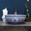 A blue and white patterned basin sits on a dark countertop, with a white sculpture and a green vase with a flower in the background. Náhled obrázku produktu