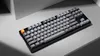 A black mechanical keyboard with gray and orange keycaps is shown on a light gray surface with a white angled background element. Produktbild-Vorschau