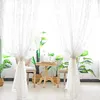 A room interior with white lace curtains, plants, and a small wooden table. Produktbild-Vorschau