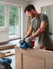 A man is using a Bosch power tool to work on a wooden door in a room with large windows. Podgląd zdjęcia produktu