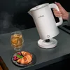 A hand is holding a white electric kettle over a base, pouring hot water into a glass of tea. A croissant with raspberries is on a plate nearby. Produktbild-Vorschau