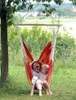 Two children are sitting in a colorful hammock chair hanging from a tree in a grassy outdoor setting. Produktbild-Vorschau