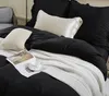 A close-up view of a neatly made bed with black bedding, a white knitted throw blanket, and decorative pillows. Produktbild-Vorschau