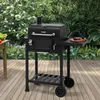 A black charcoal grill is set up on a wooden deck, surrounded by lush green foliage and flowers. Produktbild-Vorschau