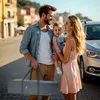A family with a baby is standing on a street with buildings in the background and a car parked nearby. The father is holding a gray travel bag. Podgląd zdjęcia produktu
