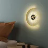 A modern wall clock with integrated lighting is mounted on a teal wall, next to a brown pillow on a bed. A small table with books and pinecones is in the foreground. Produktbild-Vorschau