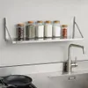 A stainless steel wall shelf mounted on a wall above a kitchen counter, holding several spice jars. A faucet and sink are visible in the foreground. Anteprima dell’immagine prodotto
