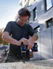 A man is drilling into a piece of wood outdoors, with a modern building in the background. Podgląd zdjęcia produktu