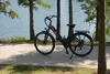 An electric bicycle is parked on a paved path next to a body of water, with trees in the foreground and background. Produktbild-Vorschau