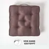 A square seat cushion with tufted details and a handle on top, set against a white background. A circular graphic with a hand sewing and text is present in the bottom left corner. Náhled obrázku produktu