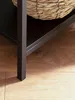 A close-up view of the leg and shelf of a dark brown console table, with a woven basket on the shelf, resting on a wooden floor. Podgląd zdjęcia produktu