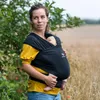 A woman is holding a baby in a black sling in an outdoor setting with trees and a field. Produktbild-Vorschau