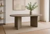 An oval dining table with a gray wood-look finish is placed on a light-colored rug in a well-lit room. A vase with green branches sits on the table. Produktbild-Vorschau