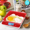 A red and white digital kitchen scale with a red bowl containing flour and an egg, with apples and a cupcake in the background. Produktbild-Vorschau