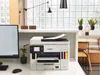 A Canon MAXIFY GX 7050 Multifunktionsdrucker is shown on a desk in a modern office setting, next to a computer monitor and a plant. The background features a window with a grid pattern and some office furniture. Produktbild-Vorschau