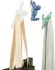 Three decorative hooks shaped like human figures are mounted on a white wall, each holding a towel or bag. Náhled obrázku produktu