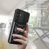 A hand holding a black phone case with a ring holder and camera cover, in a blurred indoor setting. Produktbild-Vorschau