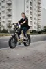 A woman rides an electric bicycle on a paved path next to a road, with a modern apartment building in the background. Produktbild-Vorschau