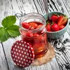 A jar of strawberries in syrup sits on a wooden table next to a bowl of fresh strawberries and a strawberry leaf. Anteprima dell’immagine prodotto